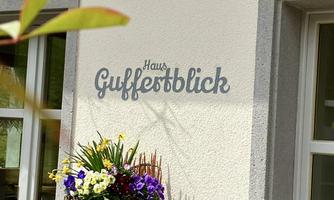 A beautifully decorated house with a sign that says "Haus Guffestblick." Colorful flowers are blooming in a pot in front of the wall.