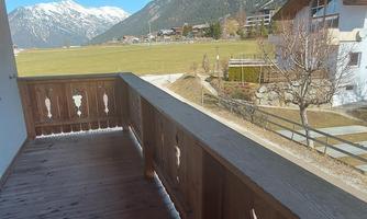 A balcony with a wooden railing and a view of green meadows and mountains. The sky is clear and blue.