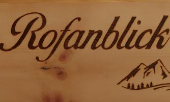 A wooden sign with the inscription "Rofanblick" and a mountain silhouette. The sign conveys a rustic and nature-oriented impression.
