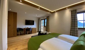 A modern hotel room with a double bed and a green bedspread. There is a television, a table with chairs, and large windows with a view of the countryside.