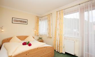 A cozy bedroom with a double bed and yellow curtains. On the bed are pillows and a small flower decoration.