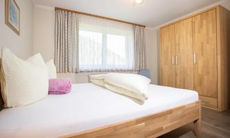 A cozy bedroom with a large bed and a light wooden wardrobe. The room has large windows and friendly curtains.
