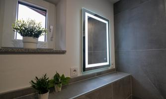 A modern bathroom with a lit mirror and plants on the windowsill. The walls are designed in neutral shades of gray.