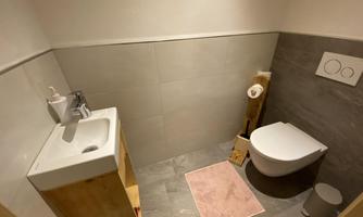 A modern bathroom with a wooden toilet bowl and sink. The floor is tiled with gray tiles and a pink bathroom rug complements the decor.