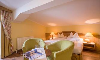 A cozy bedroom with two beds and soft yellow walls. Two green chairs and a small table create an inviting atmosphere.