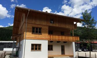 A modern wooden house with a balcony and large windows. The sky is blue with some clouds and the surroundings are green.