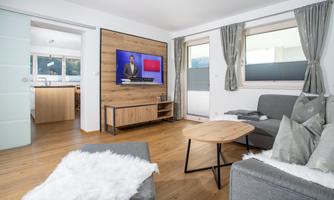 A modern living room with a gray sofa and a round wooden table. There is a television on the wall, and large windows provide plenty of natural light.