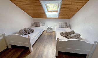 A simple bedroom with two white beds and a small nightstand. The walls are light and the ceiling is wooden, with a window for natural light.