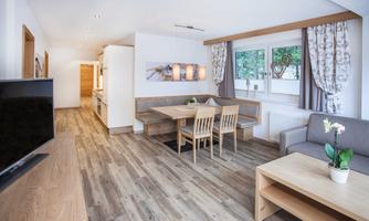 A modern living room with wooden floors, a large dining table, and a cozy seating area. Large windows provide plenty of natural light.