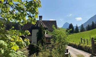 A charming house surrounded by trees and meadows. In the background, the mountains and a blue sky can be seen.