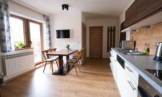 A modern kitchen with a table and chairs. Windows with curtains let in plenty of light, and a small TV is mounted on the wall.