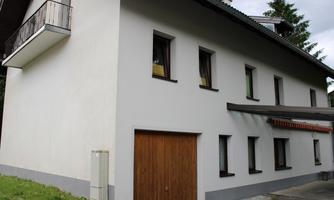 A simple, modern residential house with a bright facade and a wooden gate. It has several windows and a small balcony in the upper area.