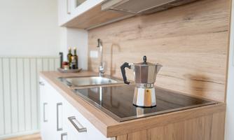 A modern kitchen with a wooden shelf and a cooking hob. On the countertop, there is a stainless steel coffee pot.