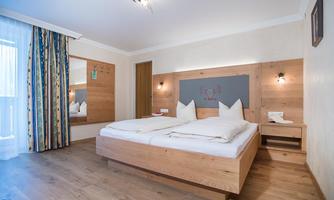 A cozy bedroom with a large double bed and wooden details. The windows are decorated with a colorful curtain and there is a mirror on the wall.