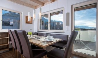 A modern dining room with a wooden table and chairs. Large windows provide a view of the mountains and the blue sky.