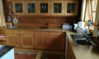A cozy wood stove kitchen with bright wooden furniture. The room is simply and functionally furnished, with windows that let in daylight.