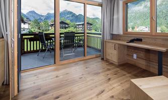 A spacious living area with large windows and a view of the mountains. The floor is made of wood, and there is a balcony with seating.