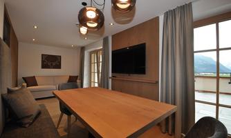 A modern living room with a large table and a cozy couch.  
Large windows provide plenty of daylight and a view of the mountains.