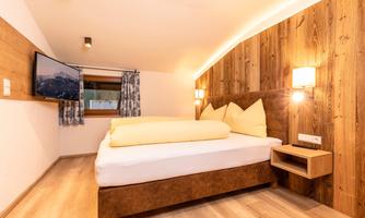 A comfortable bedroom with a large bed and wooden paneling. The room is bright and inviting, featuring a TV and windows that provide a view of the mountains.