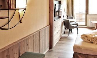 A cozy hotel room with light wood decor and a large window. There is a seating area and a modern TV console.