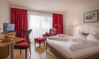 A cozy hotel room with a double bed and red curtains. Fresh fruit and a television are visible on the table.