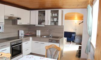 A modern kitchen with a wooden ceiling and bright furniture. In the background, a cozy living area is visible.