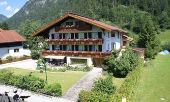 A pretty house with several floors and balconies, surrounded by green meadows and trees. The mountains are visible in the background and the sky is clear.