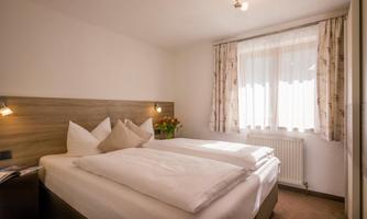 A cozy bedroom with a double bed and white bedding. The window lets in plenty of daylight and there are bright curtains.