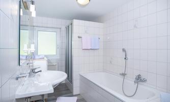 A modern bathroom with a bathtub, shower, and a sink. The walls are tiled and there is a window for natural light.