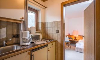 A cozy kitchen with modern appliances and a window. In the background, you can see a bright bedroom with a bed and a nightstand.
