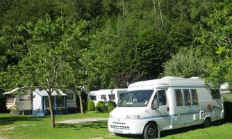 A motorhome is parked at a campsite, surrounded by trees and meadows. In the background, additional caravans are visible.