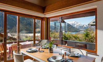 A stylish dining area with a table and dishes, surrounded by large windows. The view extends over the mountains and a clear lake.