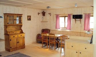 A cozy living space with wooden furniture and a table. The walls are made of light wood and there are soft curtains in pink.