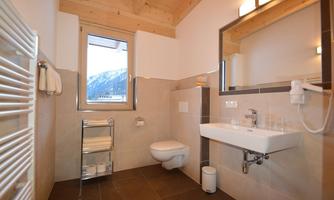 A modern bathroom with a wooden ceiling and large windows. It contains a shower, a sink, and a toilet, as well as some towels and toiletries.