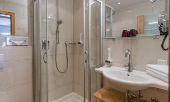 A modern bathroom with a shower cabin and a sink. The walls are light-colored tiled and there are some toiletries on the shelf.