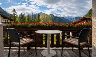 A balcony with two chairs and a table overlooks a breathtaking mountain landscape. Colorful flowers adorn the railing and the clouds drift across the sky.