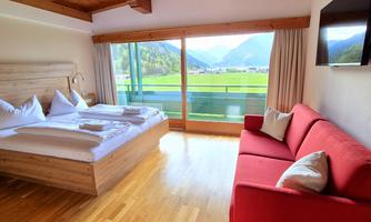 A bright hotel room with a double bed and a red couch. Large windows offer a view of the mountains and the landscape.