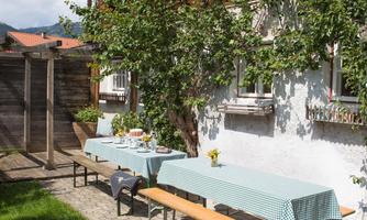 A cozy outdoor area with two tables and benches. The surroundings are green, and a typical old wooden house can be seen in the background.