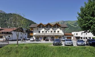 A modern building with several windows and a characteristic roof. In front of the building, several cars are parked in a parking lot, surrounded by green landscape and mountains.