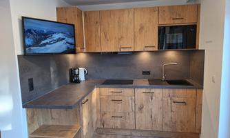 A modern kitchen with wooden cabinets and a gray countertop. A television is mounted on the wall, and there are basic kitchen appliances like a kettle.