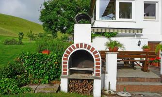 A beautiful grilling area next to a modern house, surrounded by lush greenery. In the foreground, there is a traditional stone oven and a wood rack.