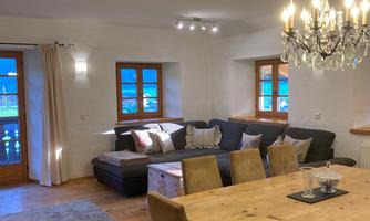 A cozy living area with a sofa, a dining table, and a beautiful chandelier. Large windows let in plenty of light and offer a view outside.