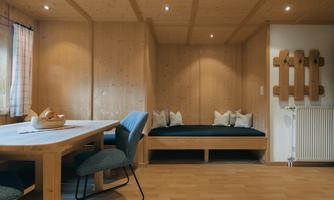 A cozy room with wooden walls and a large dining table. On a bench lie cuddly pillows and a window provides daylight.