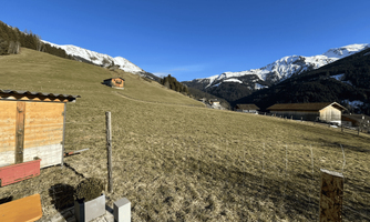 A peaceful landscape with wide meadows and snow-covered mountains in the background. In the foreground stands a wooden building, and scattered houses are present.