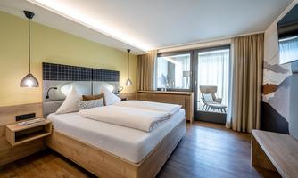 A modern hotel room with two beds and natural wood decor. Large windows provide plenty of daylight and a view outside.