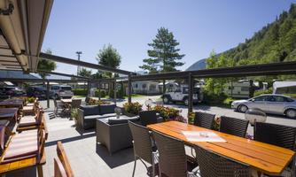 A cozy outdoor area with wooden tables and comfortable armchairs. In the background, trees and campers are visible under a clear sky.