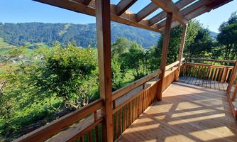 A wooden porch overlooking green hills and the clear sky. The sunlight creates beautiful shadow patterns on the floor.