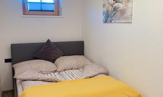 A cozy bedroom with a bed and a yellow bedspread. A painting of flowers hangs on the wall and the window lets in some daylight.
