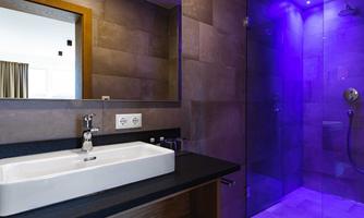 A modern bathroom with a sink and a shower. The walls are designed with dark tiles and soft, colored lighting.