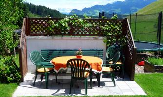 An inviting garden area with a table and chairs. In the background, green mountains and a blue sky are visible.